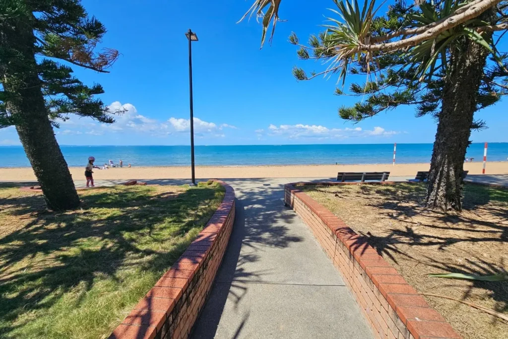 Aerial view of Suttons Beach in Redcliffe, Queensland, showing calm green waters, golden sand, high-rise buildings along the foreshore, and a clear blue sky stretching over Moreton Bay.