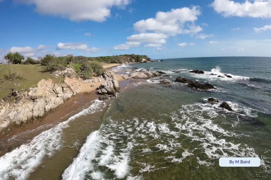 Rocky coastline and small sandy cove at Joey Leeβs Rest Beach on Curtis Island, near Gladstone, Queensland, with clear blue water, foamy waves, and green headlands under a bright sky.