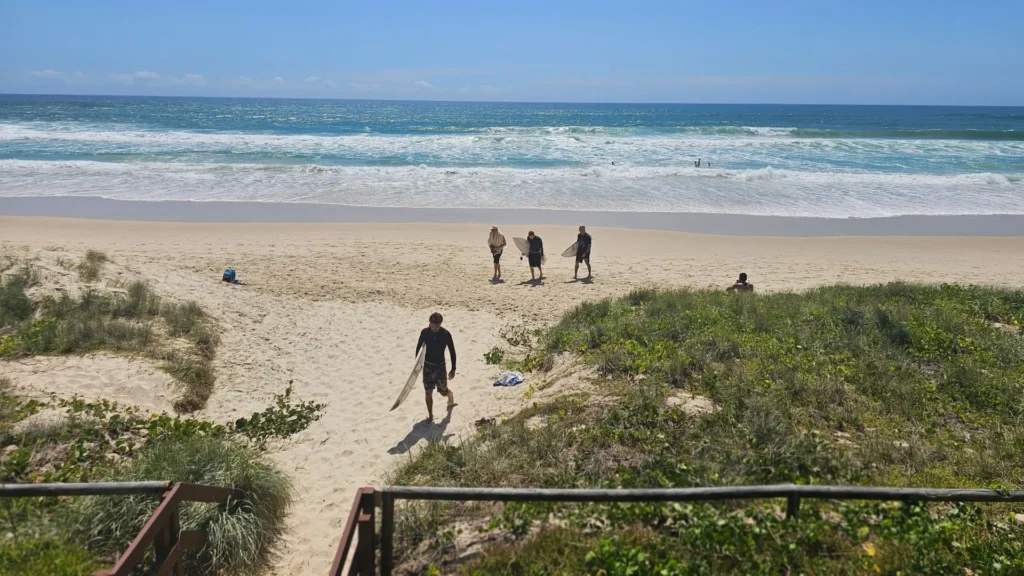 Surfers walking onto Nobby Beach Gold Coast from the dunes with waves rolling in under a clear sky