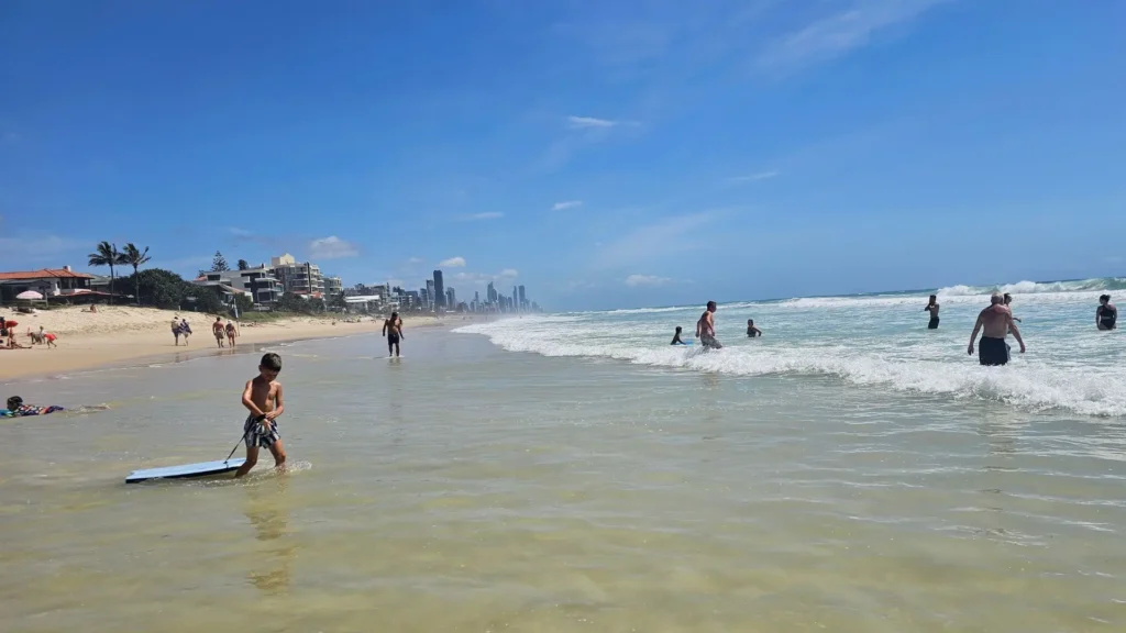 Child with a bodyboard in the shallow water at Nobby Beach with other people swimming and high-rises in the background